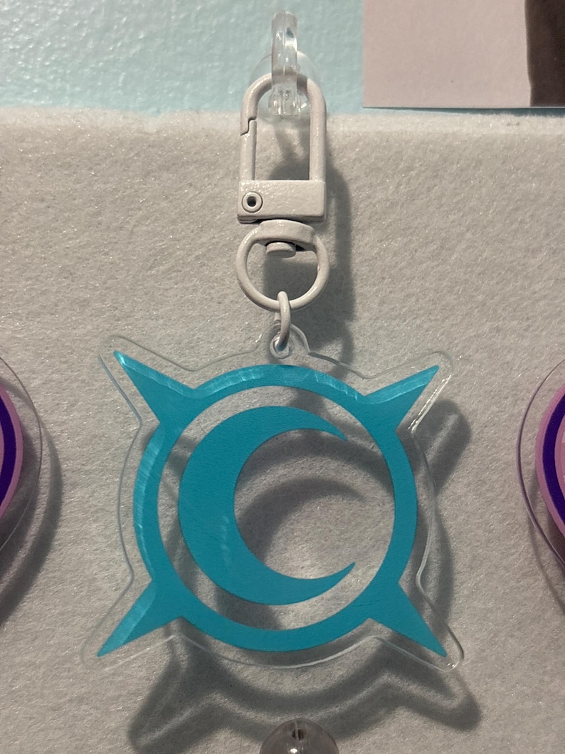 May include: A clear acrylic keychain with a light blue crescent moon and starburst design. The keychain has a silver-colored clasp for easy attachment. The design is centered on the keychain, and the overall aesthetic is modern and minimalist.