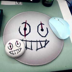 Puede incluir: Una alfombrilla de ratón gris claro con un diseño de cara de dibujos animados, con contornos negros y acentos rojos. También hay un botón blanco a juego y un ratón inalámbrico azul claro.