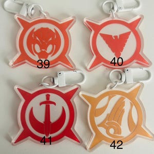 May include: Four clear acrylic keychains with white metal clasps. Each keychain features a different design in red or orange, with a white background. The designs include a snake, a bird, a sword, and an abstract shape, each with a number.