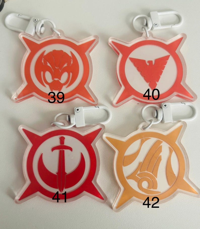May include: Four clear acrylic keychains with white metal clasps. Each keychain features a different design in red or orange, with a white background. The designs include a snake, a bird, a sword, and an abstract shape, each with a number.