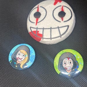 May include: A white, round, painted object with a smiling face and two smaller round buttons. The large object has a smiling face with red accents. The buttons feature a hooded figure with a face and a mouth covered with tape.