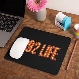 May include: Black mousepad with the text "92 LIFE" in a neon-style font. A white computer mouse sits to the left of the mousepad. A laptop, a pink flower in a vase, and office supplies are also on the wooden desk.