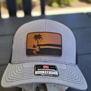 May include: A heather gray trucker hat featuring a rectangular brown leather patch with a black graphic of a palm tree, a small boat, and a shoreline. A black sticker on the brim reads EST. 1970 RICHARDSON ORIGINAL 112 TRUCKER ADJUSTABLE.