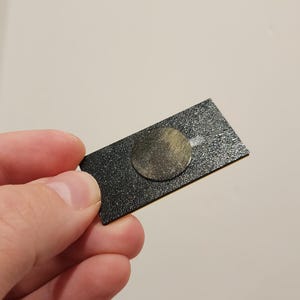 May include: A black rectangular adhesive pad with a round, gold-colored adhesive disc in the center.