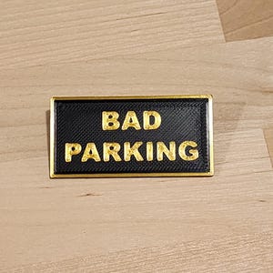 May include: A black rectangular sign with a gold border and the text "BAD PARKING" in gold letters.
