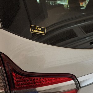 May include: A silver car with a black and yellow sticker that says "Bad Parking" on the back.