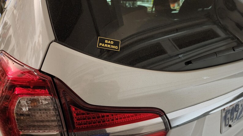 May include: A silver car with a black and yellow sticker that says "Bad Parking" on the back.