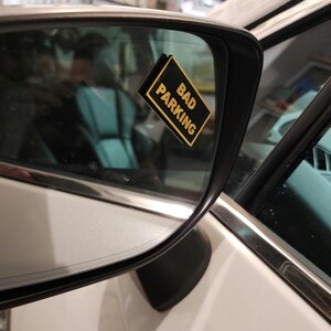 May include: A white car with a black and gold sticker that says "Bad Parking" on the side view mirror.
