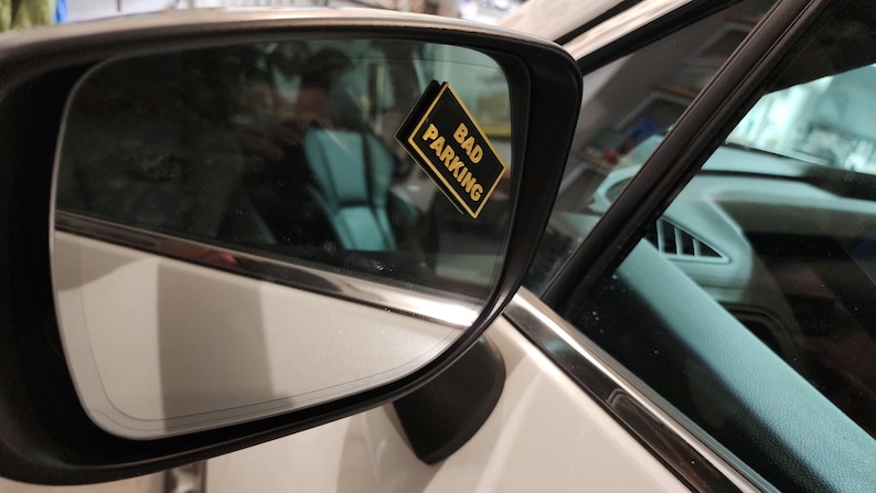 May include: A white car with a black and gold sticker that says "Bad Parking" on the side view mirror.