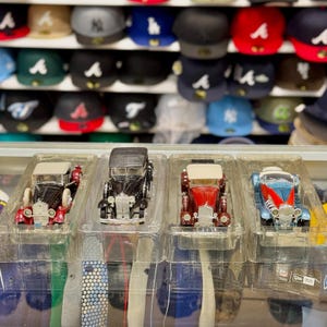 Op de afbeelding: Vier miniatuurmodellen van oldtimerauto's in doorzichtige plastic verpakking. De auto's zijn in verschillende kleuren, waaronder zwart, rood en blauw, met gedetailleerde kenmerken. Op de achtergrond is een plank met honkbalpetten te zien.