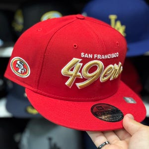 Op de afbeelding: Rode baseballcap met de woorden "SAN FRANCISCO 49ers" in goud geborduurd. De cap heeft het logo van het team aan de zijkant en een platte klep. Het is een New Era 9FIFTY-model.
