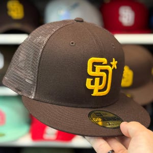 May include: A brown baseball cap with a mesh back and a flat brim. The cap features a yellow embroidered "SD" logo with a star. The New Era 59FIFTY sticker is on the brim. Other caps are visible in the background.