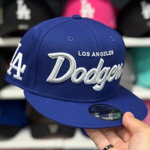 Puede incluir: Gorra de béisbol azul con el texto "LOS ANGELES Dodgers" bordado en blanco. La gorra presenta el logotipo del equipo en el lateral y una visera plana. Es un estilo ajustado.