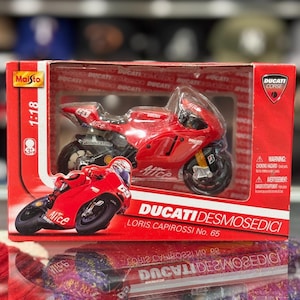 Peut inclure: Une moto jouet Ducati Desmosedici rouge dans une boîte. Le jouet est un modèle à l'échelle 1:18, avec le numéro 65 et le nom de Loris Capirossi. La boîte porte le logo Ducati Corse et un avertissement de risque d'étouffement.