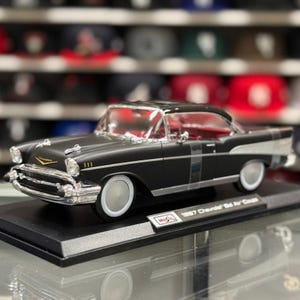 May include: A matte black 1957 Chevrolet Bel Air Coupe die-cast model car featuring chrome bumpers, trim, and white-wall tyres. The vehicle is mounted on a black display base with a plaque that reads 1957 Chevrolet Bel Air Coupe.
