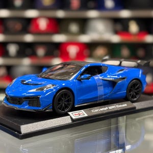 Könnte beinhalten: Blaues Druckgussmodell eines Corvette ZR1 Coupé. Das Miniaturauto hat schwarze Räder und ein schwarzes Dach. Das Modell steht auf einer schwarzen Basis mit dem Text "Corvette ZR1 Coupé".