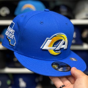 May include: A royal blue baseball cap featuring the Los Angeles Rams logo in white and yellow. The cap has a flat brim and a patch with the words "RAMS HOUSE" and a California outline. The cap is a 9FIFTY Snapback.