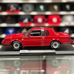 May include: A red 1987 Buick Grand National die-cast model car featuring silver wheels with BFGoodrich tires. The vehicle is mounted on a black display base with a plaque that reads Maisto 1987 Buick Grand National, viewed from the side.