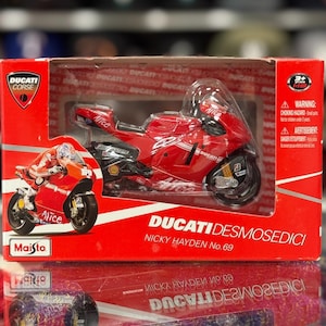 Peut inclure: Une moto jouet Ducati Desmosedici rouge dans une boîte rouge. La moto jouet a des accents noirs et blancs, avec le texte "Alice" et "Ducati" visible. La boîte indique "Nicky Hayden No. 69" et "Maisto".