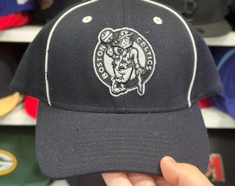 Può includere: Cappellino da baseball blu navy con il logo dei Boston Celtics. Il cappellino ha una visiera curva, profili bianchi e una corona strutturata. Il logo è un disegno ricamato di un folletto che tiene un pallone da basket.