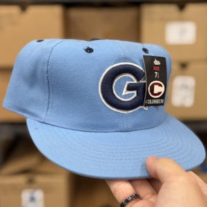 Puede incluir: Una gorra de béisbol azul claro con una letra G azul marino bordada en la parte delantera. Una etiqueta negra está unida al lateral, mostrando el texto Colosseum y la talla 7 1/2. La gorra tiene una visera plana y ojales oscuros.
