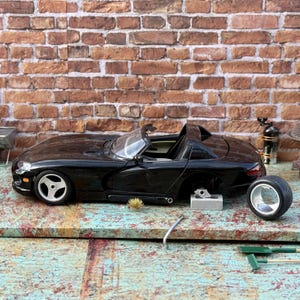 Puede incluir: Un coche modelo negro de un Dodge Viper descapotable con la capota bajada. El coche le falta una rueda y está sentado en un banco de trabajo con herramientas y un ladrillo.