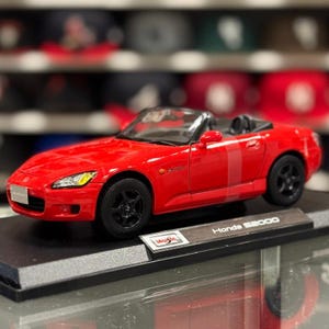 Può includere: Un modellino di auto cabriolet Honda S2000 rosso con ruote nere, fari trasparenti e interni neri. Il veicolo è montato su una base espositiva nera con il logo Maisto e la scritta Honda S2000 stampata su una targhetta color argento.