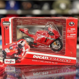 Peut inclure: Une moto jouet Ducati Desmosedici rouge dans une boîte. La moto jouet a des accents noirs et blancs et le numéro 33. La boîte présente le logo Ducati Corse et le texte "Marco Melandri No. 33". Le jouet est fabriqué par Maisto.