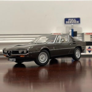 May include: A dark brown model car with silver rims and a license plate. The car is parked in front of a miniature gas station with a "FULL SERVICE" sign. The car's body has a sleek, aerodynamic design.