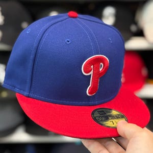 May include: A blue baseball cap with a red brim and a red "P" logo outlined in white. The cap has a red button on top and a "59FIFTY" sticker on the brim. The hat is a fitted style.