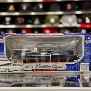 May include: A 1:18 scale die-cast model of a 1953 Cadillac Eldorado in a clear plastic display case. The car is black with silver accents. The box reads "1953 Cadillac Eldorado" and "Anson Collection".