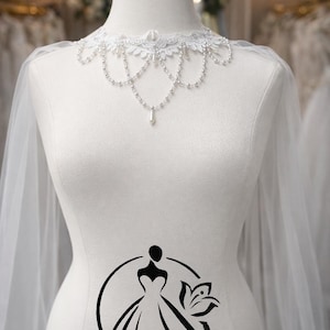 Puede incluir: Un maniquí de novia blanco exhibe un delicado cuello de encaje con detalles de perlas y un colgante en forma de lágrima. Un velo transparente cae sobre los hombros. El maniquí tiene un logotipo que dice "Hana Novias Bridal".