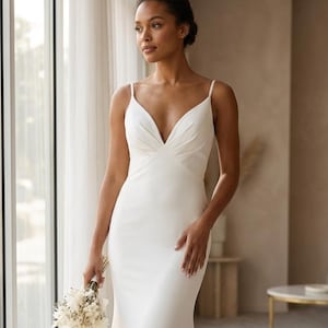May include: A white wedding dress with a V-neckline and spaghetti straps. The dress has a fitted bodice and a flowing skirt that flares out at the bottom. The bride is holding a small bouquet of white flowers.