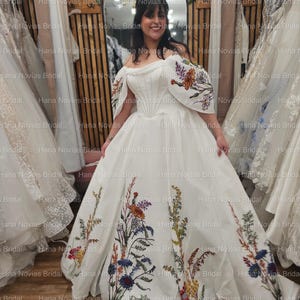 May include: A white off-the-shoulder wedding gown with floral embroidery. The dress features a fitted bodice and a full skirt adorned with colorful embroidered flowers. The sleeves also have floral embroidery. The bride is smiling in front of a mirror.