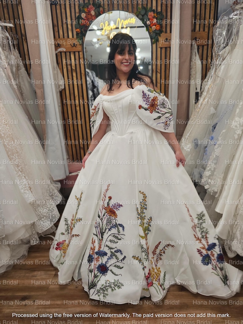 May include: A white off-the-shoulder wedding gown with floral embroidery. The dress features a fitted bodice and a full skirt adorned with colorful embroidered flowers. The sleeves also have floral embroidery. The bride is smiling in front of a mirror.