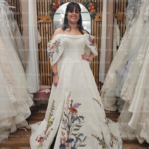 May include: A white off-the-shoulder wedding gown with floral embroidery in various colors. The dress features a fitted bodice, a full skirt, and embroidered details on the sleeves and skirt. The dress is displayed in a bridal shop.