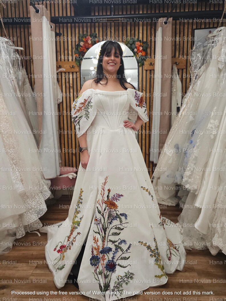 May include: A white off-the-shoulder wedding gown with floral embroidery in various colors. The dress features a fitted bodice, a full skirt, and embroidered details on the sleeves and skirt. The dress is displayed in a bridal shop.
