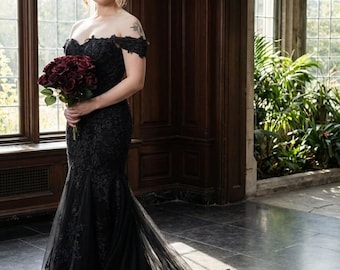 Puede incluir: Un vestido de novia negro con hombros descubiertos, corte sirena y una larga cola. El vestido presenta intrincados detalles de encaje y se combina con un ramo de flores rojo oscuro. El escenario es una habitación con grandes ventanales y paneles de madera oscura.