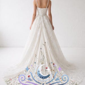 以下が含まれることがあります： 白いウェディングドレス、花の刺繍と長いトレーン付き。 ドレスは、フィットしたボディス、スパゲッティストラップ、レースのオーバーレイが特徴です。 トレーンはカラフルな花と「ENCHANTIVA」の文字で飾られています。