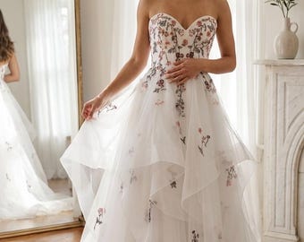May include: A strapless white wedding gown with floral embroidery. The dress features a sweetheart neckline and a layered, flowing skirt. Delicate pink and burgundy flowers are scattered across the bodice and skirt, creating a romantic and elegant look.