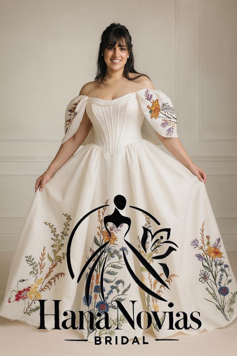 May include: Off-white bridal gown with floral embroidery. The dress features an off-the-shoulder neckline, a fitted bodice, and a full skirt. The sleeves are adorned with colorful floral designs, and the skirt has a floral pattern. The text "Hana Novias BRIDAL" is visible.