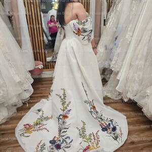 May include: A white bridal gown with an off-the-shoulder design and floral embroidery on the sleeves and train. The embroidery features a variety of colorful flowers and foliage. The dress has a flowing silhouette and a long train.