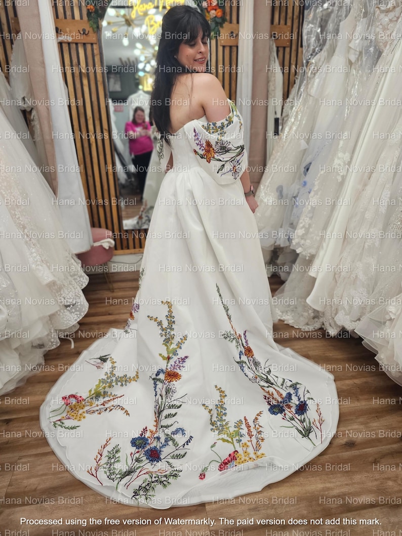 May include: A white bridal gown with an off-the-shoulder design and floral embroidery on the sleeves and train. The embroidery features a variety of colorful flowers and foliage. The dress has a flowing silhouette and a long train.