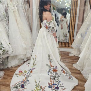 May include: A white bridal gown with floral embroidery. The off-the-shoulder dress features colorful embroidered flowers on the bodice and train. The dress is displayed in a bridal shop with other gowns in the background.