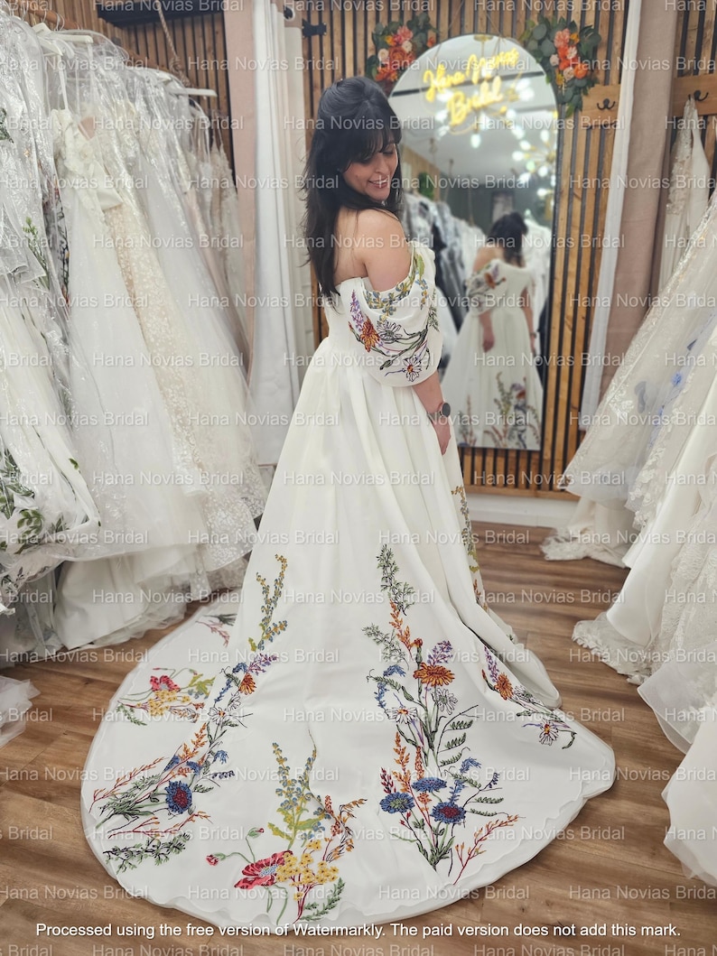 May include: A white bridal gown with floral embroidery. The off-the-shoulder dress features colorful embroidered flowers on the bodice and train. The dress is displayed in a bridal shop with other gowns in the background.