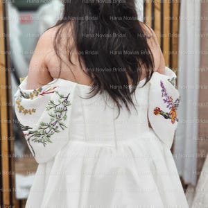 May include: A white wedding dress with floral embroidery on the sleeves. The dress has a simple, elegant design with a flowing skirt. The sleeves feature colorful embroidered flowers and greenery. The back of the dress is visible, showcasing the off-the-shoulder neckline.