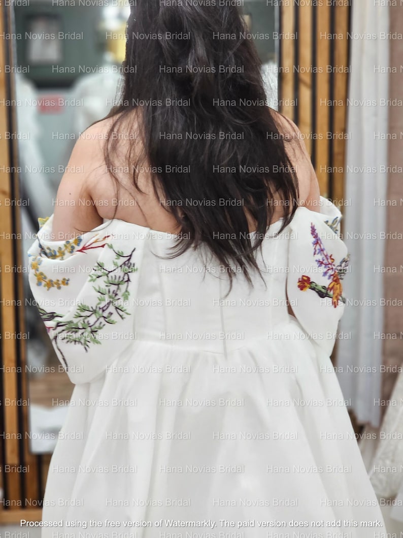 May include: A white wedding dress with floral embroidery on the sleeves. The dress has a simple, elegant design with a flowing skirt. The sleeves feature colorful embroidered flowers and greenery. The back of the dress is visible, showcasing the off-the-shoulder neckline.