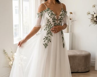 Puede incluir: Vestido de novia blanco con falda de tul fluida y delicados tirantes finos. El corpiño presenta un escote en V profundo y está adornado con detalles de hojas bordadas en verde. El vestido tiene mangas caídas y una sutil cola.