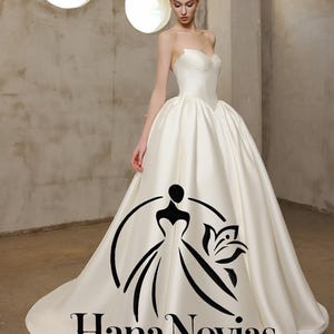 Puede incluir: Vestido de novia de satén marfil sin tirantes con falda amplia y larga cola. El vestido presenta un corpiño ajustado y una silueta de baile. El logotipo de Hana Novias Bridal está impreso en la cola.