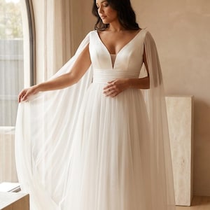 Könnte beinhalten: Ein weißes Brautkleid mit tiefem V-Ausschnitt und fließendem Rock. Das Kleid hat einen kapuzenartigen Überwurf, der über die Schultern drapiert ist. Die Braut steht in der Nähe eines Fensters, wobei natürliches Licht das Kleid hervorhebt.